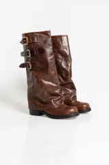 Botas de caña alta confeccionadas en cuero marrón con acabado texturizado. Presentan un diseño holgado en la caña con tres correas laterales ajustables mediante hebillas metálicas. Cuentan con puntera redondeada y suela plana de goma negra.