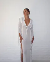 Vestido largo de microtul blanco, con escote en V profundo y mangas largas acampanadas. Presenta aberturas laterales en la falda y cierre con lazos en el frente. Es una prenda transparente.