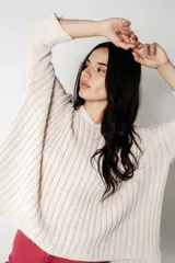 Poncho de punto color crudo con calce suelto y relajado. Confeccionado en tejido con lúrex que aporta un sutil brillo, presenta cuello en V, mangas anchas y textura acanalada.