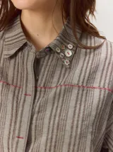 Camisa de manga corta color beige con sutil diseño a rayas verticales, cuello camisero con detalle de botones decorativos en la solapa y cierre frontal oculto.