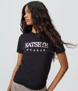 Remera negra de algodón con elastano, de corte ajustado, cuello redondo y mangas cortas. Presenta estampa frontal con la inscripción "KATSEYE" y pequeños símbolos gráficos en tono claro.
