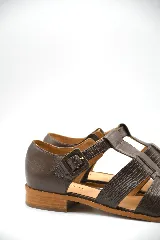 Zapato tipo cangrejera de cuero color marrón, con diseño de tiras entrelazadas, punta cerrada y pulsera con hebilla ajustable al tobillo. Cuenta con un taco bajo de madera.