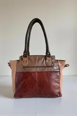 Cartera de cuero combinada en tonos marrones y beige, con detalles en animal print estilo pelo de vaca en los bolsillos frontales. Cuenta con doble asa de mano y herrajes metálicos.