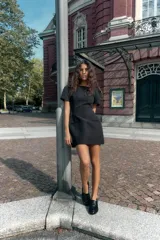 Vestido corto negro con cuello barco, mangas cortas abullonadas y pliegues. Presenta falsos bolsillos de vivo en el delantero y costuras marcadas. Cierre con cremallera oculta en la espalda.