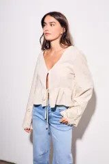 Blusa beige de viscosa y lino, con escote en V, mangas largas acampanadas con detalles bordados tipo broderie, y cierre frontal con lazo para anudar.