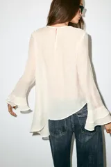 Blusa marrón de cuello redondo y manga larga, con volantes horizontales superpuestos y bajo asimétrico. Cuenta con detalle de abertura y botón en la espalda.