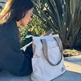 Cartera tipo tote de lona color marrón con asas de mano en color beige. Cuenta con un bolsillo frontal, bolsillos laterales y un detalle de tirador de cierre en cordón blanco.