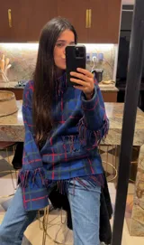 Sweater oversize de lana natural con estampado cuadrillé en tonos azul, verde y rojo. Presenta cuello alto, mangas largas y detalles de flecos torneados en el cuello, puños y dobladillo.