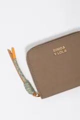 Billetera compacta de nylon color beige, con cierre de cremallera perimetral y tirador decorativo con cordón. Presenta el logo metálico de la marca en el frente.