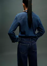 Camisa de jean con cuello camisero y lazo integrado en el frente. Presenta cierre frontal con botones, mangas largas con puños rectos y un corte recto clásico.