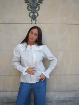 Camisa de lino color beige, con cuello bobo y detalles de encaje bordado en el cuello, pechera y puños. Cuenta con mangas largas levemente abuchonadas y cierre frontal con botones nacarados.