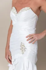Vestido de novia blanco, sin mangas, con escote corazón y espalda en V. Confeccionado en tafeta drapeada con bordados blancos. Falda con volados y tajo frontal.