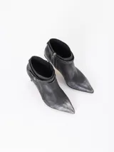 Bota corta de cuero vacuno color negro con acabado metalizado, taco fino mediano y punta fina. Presenta una tira decorativa con hebilla metálica y un dije en forma de corazón. Cuenta con cierre lateral para un calce cómodo y está completamente forrada en cuero.