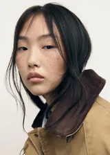 Anorak de estilo oversize en color tostado, con cuello camisero de pana en contraste. Presenta cierre frontal mediante ganchos metálicos y dos bolsillos de parche con solapa en la parte delantera.