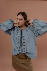 Blusa de estilo bohemio color celeste, con mangas abullonadas y amplias, escote redondo con tiras colgantes y borlas, y detalles de bordados geométricos en contraste de color en el pecho y las mangas.
