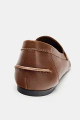 Mocasín plano de piel color marrón, estilo penny loafer con antifaz y detalle metálico dorado. Presenta punta redonda y costuras visibles.