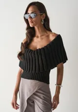 Blusa de punto negro con diseño de hombros descubiertos y escote amplio con volado acanalado. Presenta una textura de tejido acanalado vertical en toda la prenda.