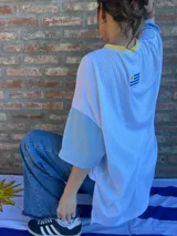 Remera de corte oversize confeccionada en tejido de punto acanalado color celeste, con mangas cortas en color amarillo y detalle de parche bordado con la bandera de Uruguay en la parte superior de la espalda.