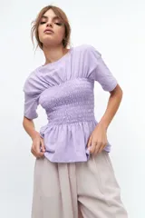 Remera de algodón color lila con cuello redondo y diseño ajustado mediante fruncido elástico tipo punto panal en la zona del busto, con mangas cortas y ruedo con volado.