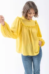 Camisa de corte holgado en color amarillo vibrante, confeccionada en tela fluida. Presenta cuello alto con volados, detalle de frunces en el canesú y mangas largas abullonadas con puños elásticos.