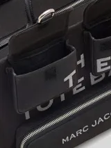 Cartera tote de lona color negro, modelo The Cargo Canvas Medium Tote Bag de Marc Jacobs. Presenta múltiples bolsillos frontales con solapa y cierre de cremallera, asas de mano superiores y una correa de hombro ajustable y extraíble. Cuenta con el nombre de la marca y la inscripción "THE TOTE BAG" estampados en el frente.