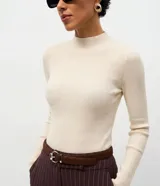 Suéter de punto canelado color beige, con cuello alto y manga larga. Presenta un diseño ajustado al cuerpo, confeccionado en una mezcla de viscosa, poliéster y poliamida.