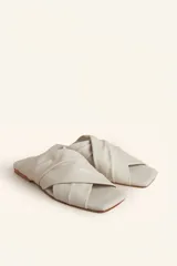 Sandalia tipo slide de cuero vacuno color blanco, con diseño de tiras cruzadas y fruncidas. Presenta una puntera cuadrada y una suela plana de cuero con un pequeño taco de 2 cm.
