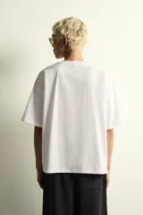 Camiseta de corte holgado con cuello redondo y manga corta. Presenta un diseño frontal con la frase New York Essence combinando tipografía cursiva y letras en bloque con efecto relieve.