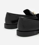 Zapato mocasín femenino de PU color negro, con punta cuadrada y detalle metálico dorado en el empeine. Tiene un tacón bajo y suela de goma.