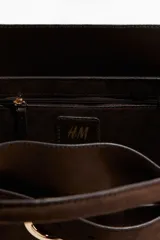 Bolso tote de acabado suave, color marrón oscuro, con asas tubulares y detalle de correa frontal con hebilla metálica dorada.