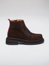 Botineta de gamuza marrón con detalles en cuero liso al tono. Presenta una suela track negra con costuras en contraste color rojo y una tira vertical roja en el talón.