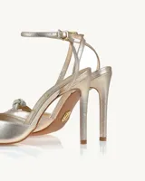 Zapato de tacón alto color dorado con pulsera al tobillo y detalle de moño plateado con brillos en la punta.
