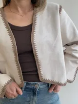 Chaqueta de efecto gamuza color tostado, con diseño abierto sin cierres y detalles de costuras artesanales tipo crochet en color blanco en los bordes, el frente, el cuello y las mangas.