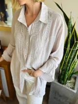 Camisa de manga larga en color visón claro, con diseño de tablitas verticales y detalles de puntilla. Presenta cierre frontal con botones de nácar y cuello camisero clásico.