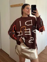 Sweater de punto color marrón con diseño oversize, cuello redondo y mangas largas. Presenta bordados artesanales en relieve con hilos de colores y detalles en lurex, incluyendo flecos decorativos distribuidos por toda la prenda.