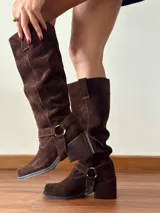 Botas de caña alta confeccionadas en cuero gamuzado color chocolate. Presentan un diseño de estilo western con puntera cuadrada, taco bloque de altura media y un estribo desmontable con argolla metálica en la zona del tobillo.