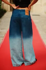 Jean de tiro medio con corte oversize y botamanga ancha. Presenta un diseño de lavado degradado que va de un azul intenso en la cintura a un tono claro en la parte inferior, con efecto gastado. Incluye avíos metálicos y badana de PU.