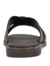 Sandalia plana color marrón oscuro, confeccionada en cuero, con tiras cruzadas en el empeine y tachas metálicas doradas decorativas.