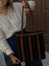 Cartera tipo tote de cuero con diseño de franjas verticales alternadas en tonos negro y marrón. Cuenta con manijas trenzadas de cuero y cierre superior.