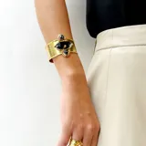 Brazalete rígido tipo cuff realizado en bronce con acabado texturizado. Cuenta con una piedra natural central de forma geométrica en color gris y dos piedras naturales laterales en forma de gota de color rojo. Diseño ajustable de 3 cm de ancho.