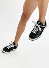 Zapatillas urbanas negras con detalles en cuero, gamuza y charol. Presentan cordones plateados, suela color crema y logo "C" en el lateral.