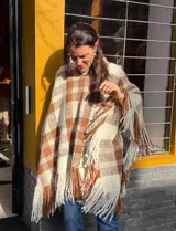 Poncho de lana merino tejido en telar con diseño de cuadros en tonos beige, marrón y blanco, con terminación de flecos largos en los bordes.