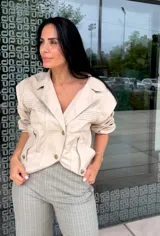 Chaqueta color nude con cuello solapa, cierre frontal de botones dorados y bolsillos laterales. Presenta detalles de pliegues horizontales en los hombros y verticales en la cintura.