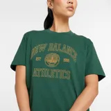 Remera New Balance de corte oversize en color verde oscuro, de manga corta y cuello redondo. Presenta un estampado frontal con tipografía y un diseño circular de escudo conmemorativo.