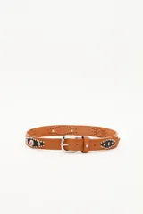 Cinturón de cuero vacuno color suela con hebilla metálica plateada grabada. Presenta apliques de cuero negro con tachas metálicas y piedras decorativas de colores.