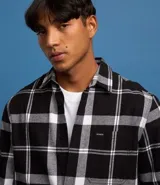 Camisa de franela de manga larga con estampado de cuadros en blanco y negro. Presenta cuello inglés, cierre frontal con botones y bolsillo en el pecho.