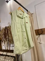 Musculosa tipo camisa con cuello camisero, cierre frontal de botones y dos bolsillos delanteros en el pecho. Diseño a rayas verticales en tonos celeste y blanco.