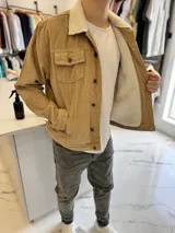 Chaqueta de pana color marrón tostado, con forro interno y cuello de corderito sintético blanco. Presenta cierre frontal de botones metálicos, bolsillos frontales con solapa en el pecho y bolsillos laterales.