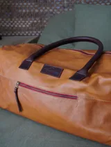 Bolso tipo weekender de cuero marrón, con asas de mano y bolsillo frontal con cierre metálico.