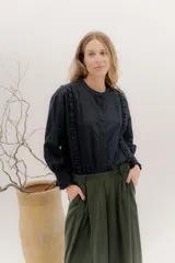 Camisa de algodón blanca con cuello a la base, cierre frontal con botones y mangas largas con puños delicados. Presenta volados verticales y detalles de cintas de terciopelo en contraste a lo largo del frente.